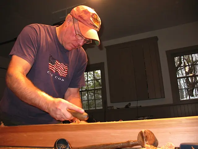 Man wearing a cap and glasses working with wood indoors.